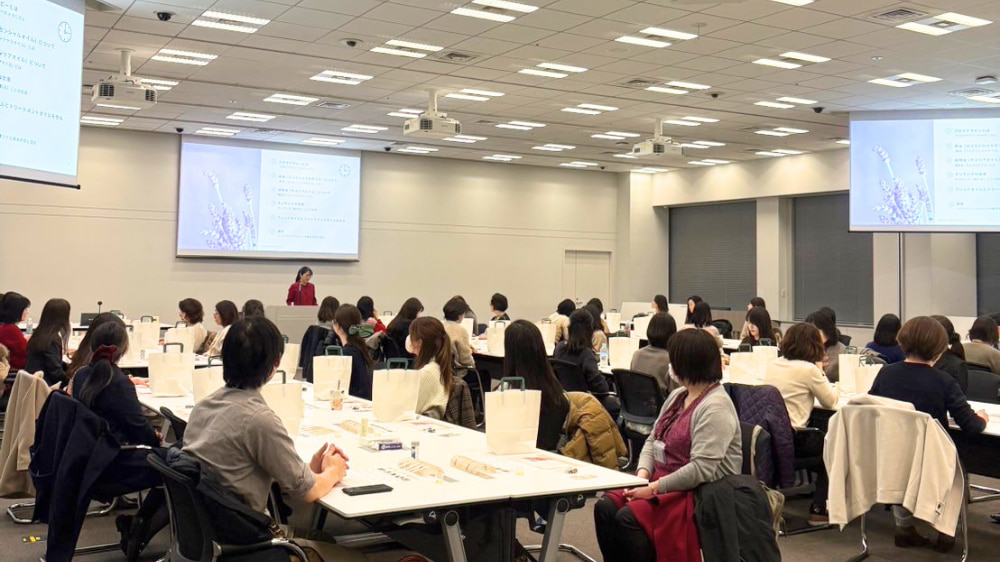 企業向けセミナーでアロマテラピーについて講義する講師の様子。参加者に向けてプロジェクターを使いながら説明している場面。